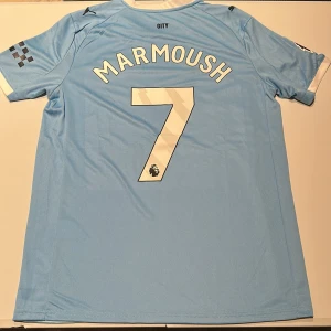 Manchester City Marmoush #7 fotbollströja - Snygg Manchester City fotbollströja från Puma med Marmoush och nummer 7 på ryggen. Tröjan är ljusblå med vita och mörkblå detaljer, har korta ärmar och diagonal randig design framtill. Officiellt klubbmärke och Premier League-logga. Tillverkad i lätt och ventilerande polyester. Replika, endast handtvätt gäller!