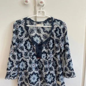 Mönstrad blus från Odd Molly, blå/vit - Supersöt blus från Odd Molly i blått och vitt med unikt mönster. Blusen har v-ringning med spetsdetaljer, knappar framtill och trekvartsärmar med lätt volang. Tillverkad i tunn bomull, perfekt för vår och sommar. 