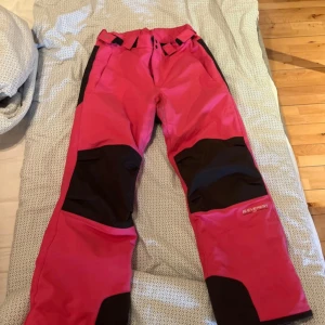 Rosa skidbyxor från Everest - Säljer ett par snygga rosa skidbyxor från Everest med svarta förstärkningar på knän och benslut. Byxorna har justerbar midja med kardborre, bälteshällor och dragkedja fram. Perfekta för vinterns alla äventyr i snön!