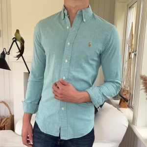 Ljusblå skjorta från Ralph Lauren - Snygg ljusblå skjorta från Ralph Lauren med klassisk button-down krage och broderad logga på bröstet. Skjortan är i bomull, har lång ärm och normal passform. Perfekt till jeans för en clean och stilren look.