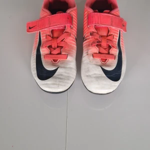 Nike fotbollsskor i rött och vit 36 - Snygga fotbollsskor från Nike i rött och vitt med svarta detaljer och stor svart swoosh på sidan. Skorna har snörning och kardborreband för extra bra passform samt dobbar för gräsplan. Perfekta för dig som vill sticka ut på planen.
