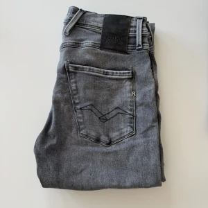 Replay Anbass grå jeans, storlek 30/32 - Snygga Replay Anbass jeans i grå tvätt med klassisk femficksdesign och svarta detaljer. Modellen är slim och har raka ben, tillverkad i stretchigt jeansmaterial för skön passform. Perfekta för dig som gillar en modern och stilren look.