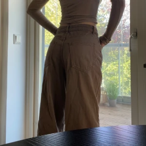 Bruna wide jeans med hög midja - Säljer ett par bruna jeans med hög midja och breda ben. Byxorna har klassisk femficksdesign, raka sömmar och stängs med knapp och dragkedja. Perfekta för dig som gillar en avslappnad och trendig look. Helt nya! För stora för mig.