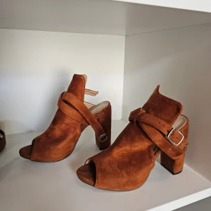 Bruna mockasandaler med klack - Snygga sandaler i brun mocka med öppen tå och bred klack. Skorna har en cool omlottrem med spänne runt vristen och en stabil plattform. Perfekta för dig som vill ha en trendig och bekväm sko med lite höjd.