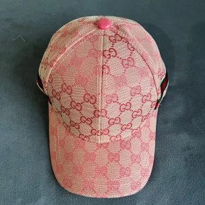 Gucci rosa keps med GG-mönster - Snygg rosa keps från Gucci med klassiskt GG-mönster i ljusrosa och rosa toner. Kepsen har justerbar rem baktill i rosa läder och detaljer med grönt och rött band på sidan. Tillverkad i bomull och polyester, perfekt för dig som vill sticka ut med lyxig streetstyle.