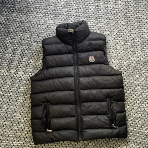 Byter - Snygg svart dunväst från Moncler med hög krage och dragkedja framtill. Västen har två fickor med dragkedja och klassisk Moncler-logga på bröstet. Perfekt för lager-på-lager och ger en clean, sportig look.