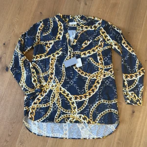 Helt ny damskjorta från Stenströms - Snygg henley-skjorta från Stenströms med marinblå botten och guldiga kedjemönster. Skjortan har v-ringning, knappar framtill, två bröstfickor och långa ärmar. Materialet känns lätt och svalt. Oanvänd med etiketter kvar. Storlek dam 40, motsvarar medium till large.