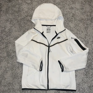 Nike tech - Säljer en Vit Nike tech med svarta detaljer och dragkedjor. Tröjan har huva, lång ärm och en extra ficka med dragkedja på ena ärmen. Snygg kontrast med svarta kanter och Nike-logga på bröstet. Perfekt för dig som gillar sportig och clean stil.