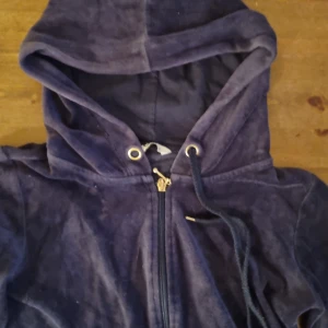 Mörkblå velour hoodie med dragkedja - Mysig mörkblå hoodie i velour med huva och snörning. Tröjan har dragkedja framtill, fickor och guldfärgade detaljer vid snörningen. Perfekt för chill dagar eller när du vill vara extra bekväm.