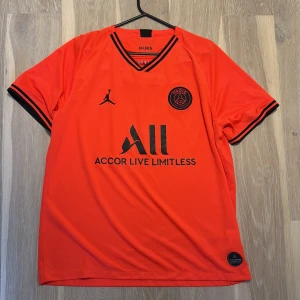 PSG x Jordan röd matchtröja - Snygg PSG fotbollströja i orange med svarta detaljer från Jordan. Kortärmad modell med svart logga och klubbmärke på bröstet, samt svart text och ränder på ärmsluten. Tillverkad i lätt och ventilerande polyester, perfekt för träning eller match. Köptes för 2 år sen, knappt använd.