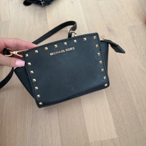Svart axelväska Michael Kors med nitar - Snygg svart axelväska från Michael Kors med guldfärgade nitar runt kanterna och logga framtill. Väskan är i skinn och har justerbar axelrem samt dragkedja upptill. Perfekt storlek för det viktigaste och ger en cool vibe till din outfit.