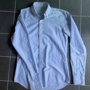 Blåvit randig skjorta John Henric - Snygg blå och vit randig skjorta från John Henric med klassisk krage och knappar framtill. Skjortan är långärmad och tillverkad i bomull, perfekt för en clean och fräsch look. Passformen är normal och materialet är lätt att stryka. Storlek 37, sitter som S!