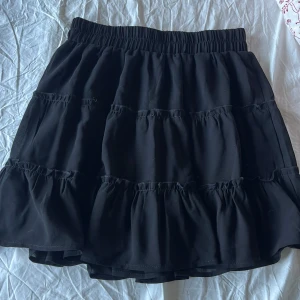 Svart volangkjol med resårmidja - Supersöt svart kortkjol med flera volanglager och bekväm resår i midjan. Kjolen har en luftig känsla och passar perfekt till sneakers eller boots. Enkel att styla och ger en trendig look.