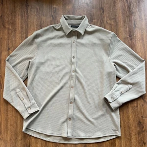 Beige skjorta från Brand Island XXL - Stilren beige skjorta från Brand Island i storlek XXL. Skjortan har lång ärm, klassisk krage och knäppning framtill med snygga träfärgade knappar. Materialet är strukturerat och känns mjukt, perfekt för en clean och avslappnad look.