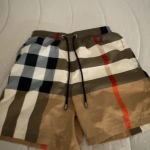 Rutig Burberry shorts med snörning - Snygga Burberry shorts med klassiskt rutigt mönster i beige, svart, vit och röd. De har elastisk midja med svart snörning och meshfoder på insidan. Perfekt för sommaren och chill dagar. Bakficka med rutigt tyg för extra stilpoäng.