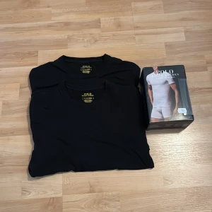 Ralph lauren 2 pack t-shirts - Svart t-shirt från Polo Ralph Lauren i bomull med klassisk passform. T-shirten har korta ärmar, rund halsringning och en röd broderad logga vid nederkanten. Perfekt basic-plagg med stilren look. (helt oanvända)