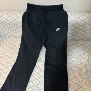 Svarta mjukisbyxor från Nike - Svarta mjukisbyxor från Nike med dragsko i midjan och broderad vit Nike-logga på benet. Byxorna har bakficka med röd Nike-tag och är gjorda i mjukt bomullsmaterial. Perfekta för chill eller träning.