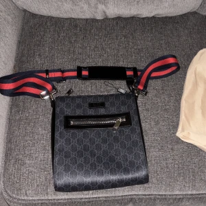 Gucci axelväska med GG-mönster  - Snygg axelväska från Gucci i mörkgrått med klassiskt GG-mönster. Väskan har en bred, justerbar axelrem i rött och marinblått, samt detaljer i svart skinn. Framsidan har en praktisk dragkedjeficka och väskan stängs med dragkedja upptill.