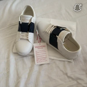 Valentino Garavani vita sneakers - Säljer ett par snygga vita Valentino Garavani sneakers i skinn med bred mörkblå rem över vristen och vita snören. Klassisk rund tå och platt sula med nitar baktill. Kom privat för fler bilder, helt nya använda 1gång.