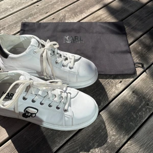 Vita sneakers från Karl Lagerfeld - Snygga vita sneakers från Karl Lagerfeld med svart häl och ikonisk Karl-figur på sidan. Skorna har snörning, rund tå och platt sula. Tillverkade i skinn och levereras med originalpåse. Perfekta för dig som gillar stilrena och trendiga skor. Nypris ca 2300kr