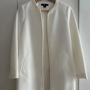 Vit kappa från H&M - Säljer en stilren vit kappa från H&M med rak passform och diskret struktur i tyget. Jackan har lång ärm, två fickor framtill och är helt utan knappar eller dragkedja för en clean look. Perfekt för dig som gillar minimalistisk stil