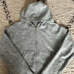 Grå stickad hoodie från H&M - Mysig grå hoodie från H&M i stickat material med dragkedja framtill och huva. Tröjan har långa ärmar, ribbade muddar och två praktiska fickor. Perfekt för kyliga dagar när du vill vara både bekväm och stilren.