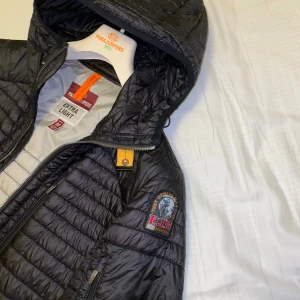 Svart lättviktsjacka Parajumpers - Svart quiltad lättviktsjacka från Parajumpers med huva, dragkedja och ikonisk patch på ärmen. Jackan har en gul detalj vid halsen och flera praktiska fickor. Perfekt för dig som vill ha en snygg och funktionell jacka med sportig vibe.