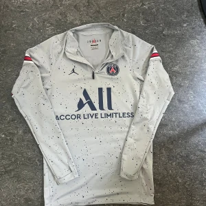 PSG x Jordan långärmad tröja  - Säljer en grå långärmad PSG x Jordan fotbollströja i slim fit med svart prickigt mönster. Tröjan har halv dragkedja fram, PSG-logga på bröstet och Jordan-logga. Röda och svarta ränder på ärmarna. Materialet är mjukt och stretchigt, perfekt för träning eller vardaglig använding