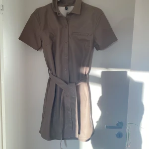 Beige kort klänning i skinnimitation Storlek S - Snygg beige kort klänning från H&M i skinnimitation med korta ärmar, knappar hela vägen framtill och ett matchande bälte i midjan. Stilren design med liten krage och bröstficka, perfekt för dig som gillar minimalistisk look.
