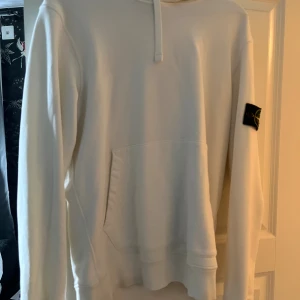 Vit hoodie från Stone Island - Snygg vit hoodie från Stone Island med klassisk patch på ärmen. Tröjan har huva med snörning och en stor magficka framtill. Perfekt för dig som gillar stilrena och exklusiva streetwear-plagg. Köpt från NK i Göteborg för runt 3000. Storlek L.