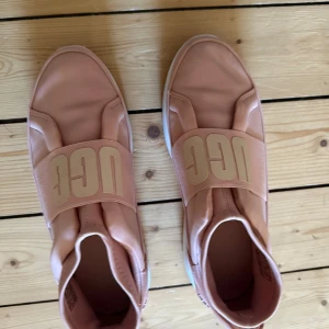 Rosa sneakers från UGG med bred resår - Snygga sneakers från UGG i ljusrosa med bred resår över foten och stor UGG-logga. Ovandel i syntet och mockadetaljer, vit platt sula och rund tå. Perfekta för dig som gillar sportig och clean stil.