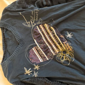 Svart långärmad tröja med broderi - Unik svart långärmad tröja med broderat hjärta och blommor framtill. Ärmarna har coola mönster och ribbade muddar i grått. Tröjan har v-ringning och detaljer i flera färger, perfekt för dig som gillar personliga plagg med lite edge.