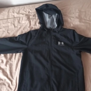 Under armour vindjacka bra condition och allting på den är helt bra. - Svart vindjacka från Under Armour med huva och hel dragkedja. Jackan har meshfoder på insidan för extra ventilation och en stilren logga på bröstet. Perfekt för blåsiga dagar och sportiga outfits.