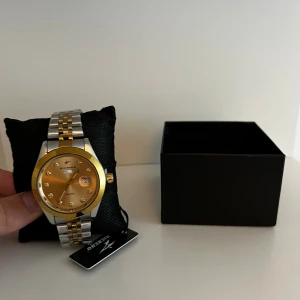 Klocka  - ✨ Klassisk och elegant klocka från Ricecgo 	•	Helt ny och oanvänd 	•	Tvåfärgad design i silver och guld 	•	Champagnefärgad urtavla med gnistrande stenar som timmarkörer 	•	Quartz urverk – pålitligt och exakt 	•	Justerbart armband i slitstark metall  En tidlös modell som ger en exklusiv känsla till varje outfit.