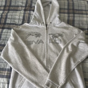 Grå hoodie med BVALDI tryck och strass - Säljer en grå hoodie från BVALDI med strassdetaljer och coolt tryck av två händer på bröstet. Tröjan har huva, dragkedja och långa ärmar. Perfekt för dig som gillar streetstyle och vill sticka ut med snygga detaljer. 