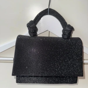 Svart glittrig handväska med knutdetaljer - Snygg svart handväska med glittriga paljetter över hela väskan och coola knutdetaljer på handtaget. Väskan har en stilren, rektangulär form och passar perfekt när du vill addera lite extra bling till din outfit. Handtaget är robust och dekorerat med små glittrande stenar.