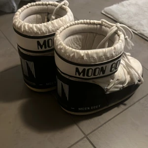Svarta och vita Moon Boots - Säljer ett par klassiska Moon Boots i svart och vitt med tydlig logga runt skaftet. Skorna har snörning upptill och är vadderade för extra värme. Perfekta för vintern och riktigt sköna att ha på sig när det är kallt ute.