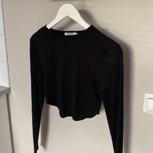 Svart croppad långärmad topp från NA-KD - Svart ribbad långärmad topp från NA-KD i croppad modell, storlek S. Enkel och stilren med rund halsringning, perfekt att matcha med höga jeans eller kjol. Tillverkad i mjuk och stretchig polyester för en bekväm passform.