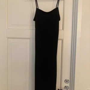 Svart långklänning med smala axelband - En stilren svart långklänning med smala axelband och rak passform. Klänningen är tillverkad i mjuk och stretchig polyesterblandning som ger en snygg siluett. Perfekt för dig som gillar minimalistisk och tidlös stil.
