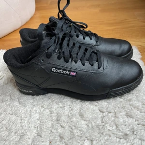 Svarta Reebok sneakers storlek 39 - Klassiska svarta Reebok sneakers i skinn med låg profil och rund tå. Skorna har svart snörning, diskret Reebok-logga på sidan och bak, samt en platt sula. Perfekta för en clean och stilren look. Använda vid två tillfällen. 