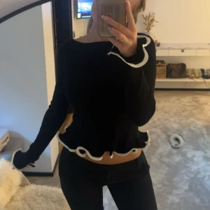 Svart tröja med volangkant - Supersöt svart tröja med vita vågiga volangdetaljer vid ärmslut och nederkant. Tröjan har lång ärm och är lite cropped. 