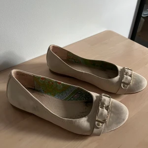Beige ballerinaskor med gulddetaljer - Snygga beigea ballerinaskor med rund tå och dekorativ guldfärgad spänne framtill. Skorna har ett mjukt skinnliknande material och färgglatt mönstrat innerfoder. Har lite fläckar utanpå på sidan och i skorna men fortfarande i bra skick.