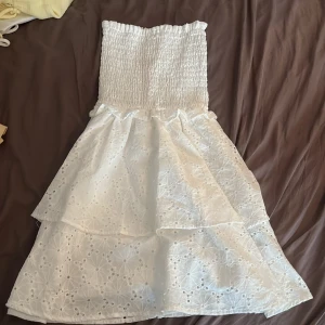 Vit  klänning  - Supersöt vit klänning med smockad överdel och volangig kjol. Ärmlös och axelbandslös modell, perfekt för varma dagar. Blommigt hålmönster ger en romantisk vibe, perfekt för skolavslutning, och kjolen har två lager för extra volym. Helt alldeles ny, aldrig använd