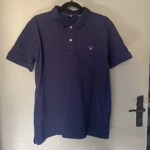 Marinblå pikétröja från GANT - Snygg marinblå pikétröja från GANT med klassisk krage och två knappar framtill. T-shirten har korta ärmar och en liten broderad logga på bröstet. Tillverkad i mjuk bomull som är skön mot huden. Perfekt för en chill och stilren look.