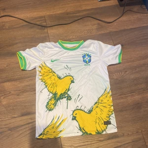 Vit Brasilien fotbollströja Nike M - Snygg vit Brasilien fotbollströja från Nike med gröna och gula detaljer på krage och ärmslut. Framsidan har stora gula fåglar och det klassiska CBF-emblemet på bröstet. Tillverkad i Dri-FIT-material som andas och passar perfekt för match eller träning. Står S men passar som M, använd typ 5 gånger som ny 