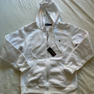 Vit hoodie från Polo Ralph Lauren - Vit hoodie från Polo Ralph Lauren med dragkedja, huva med snörning och klassisk broderad logga på bröstet. Tröjan har känguruficka framtill och ribbade muddar vid ärmslut och nederkant. Perfekt för en clean och stilren look.