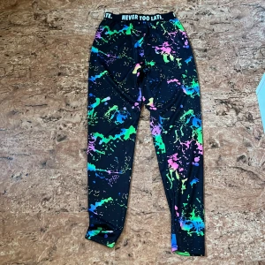 Färgstarka leggings med tryck, 10-11 år - Leggings med svart bas och färgstarkt stänkmönster i grönt, blått, rosa och gult. Resårmidja med texten 'NEVER TOO LATE' runtom. Tillverkade i stretchigt syntetmaterial, perfekta för träning eller chill. Sitter tajt och följer kroppen.