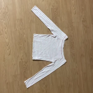 Vit och rosa randig långärmad topp Kappahl - Vit långärmad topp från Kappahl med tunna rosa ränder. Tröjan har en lätt ribbad struktur och små volangdetaljer vid ärmslut och nederkant. Perfekt för dig som gillar en söt och enkel stil.