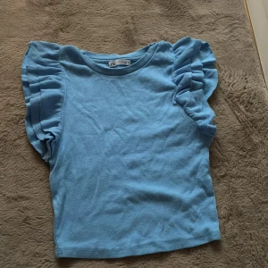 Blå volangtopp från Zara - Supersöt blå topp från Zara med volangdetaljer på axlarna. Toppen är ribbad och har en rund halsringning. Perfekt för dig som vill ha något som sticker ut lite extra i garderoben. Storlek M men passar mig perfekt som har storlek S. Använd en gång så som nyskick 