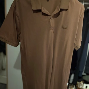 Brun pikétröja från Lacoste - Säljer en brun pikétröja från Lacoste med klassisk krage och knappslå. Tröjan har korta ärmar och den ikoniska krokodilloggan broderad på bröstet. Perfekt för en stilren och avslappnad look.